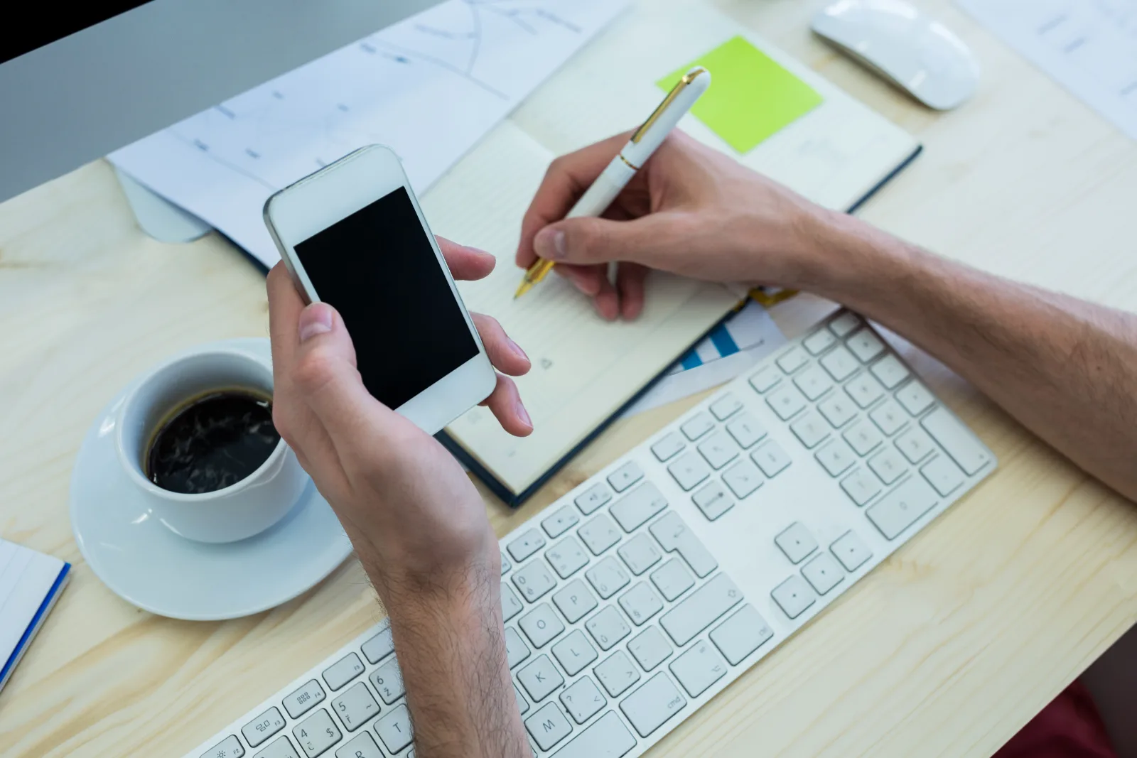 Hands writing in a notebook while holding a phone at a desk.